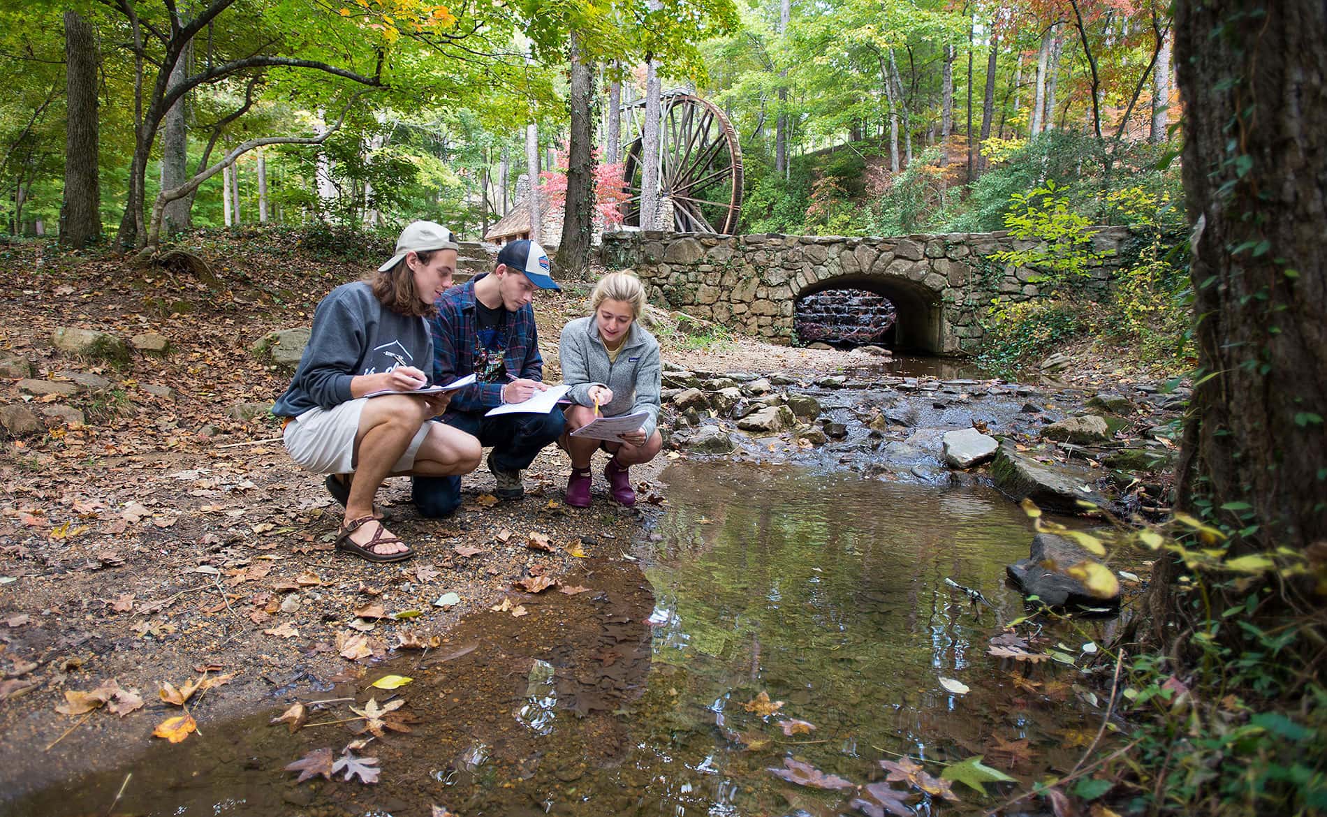 three students kneeling by a creek examinig papers
