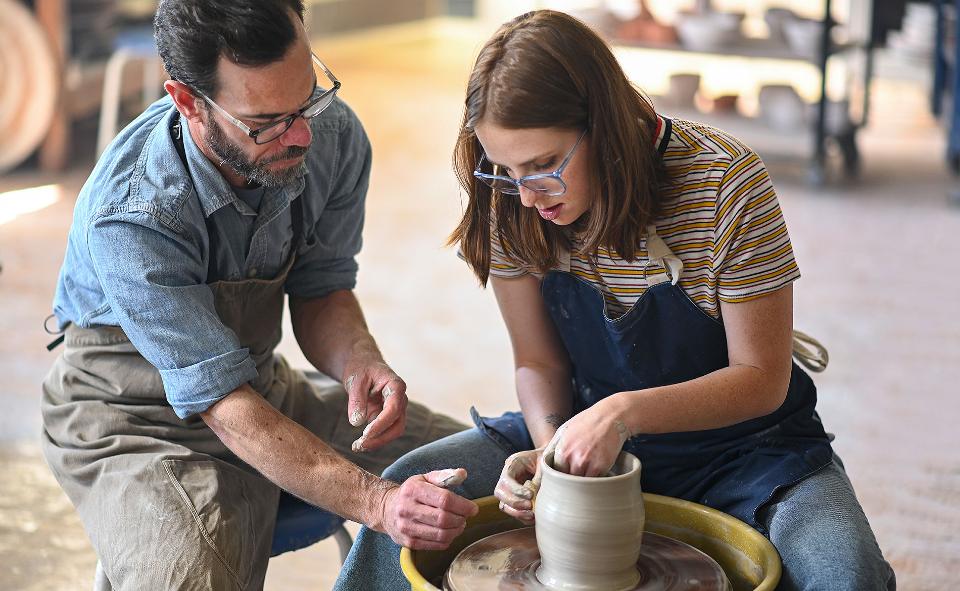 professor kneeling by student making a clay vase