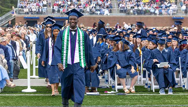 Male graduate leading a line of other graduates to the stage at Commencement