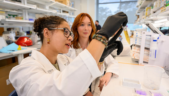 A female student and a professor in lab coats observing liquid in a tube