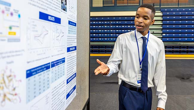 Male student speaking and pointing at a large poster containing text and data graphs