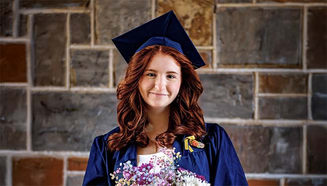 Nancy Daniel, smiling and holding a bouquet, standing in front of a brick wall, wearing a graduation cap and gown