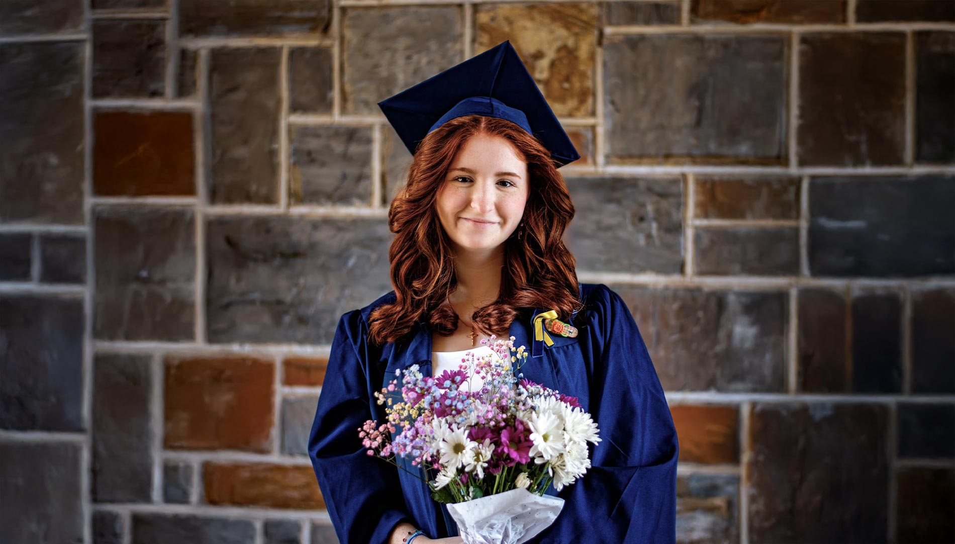 Nancy Daniel, smiling and holding a bouquet, standing in front of a brick wall, wearing a graduation cap and gown